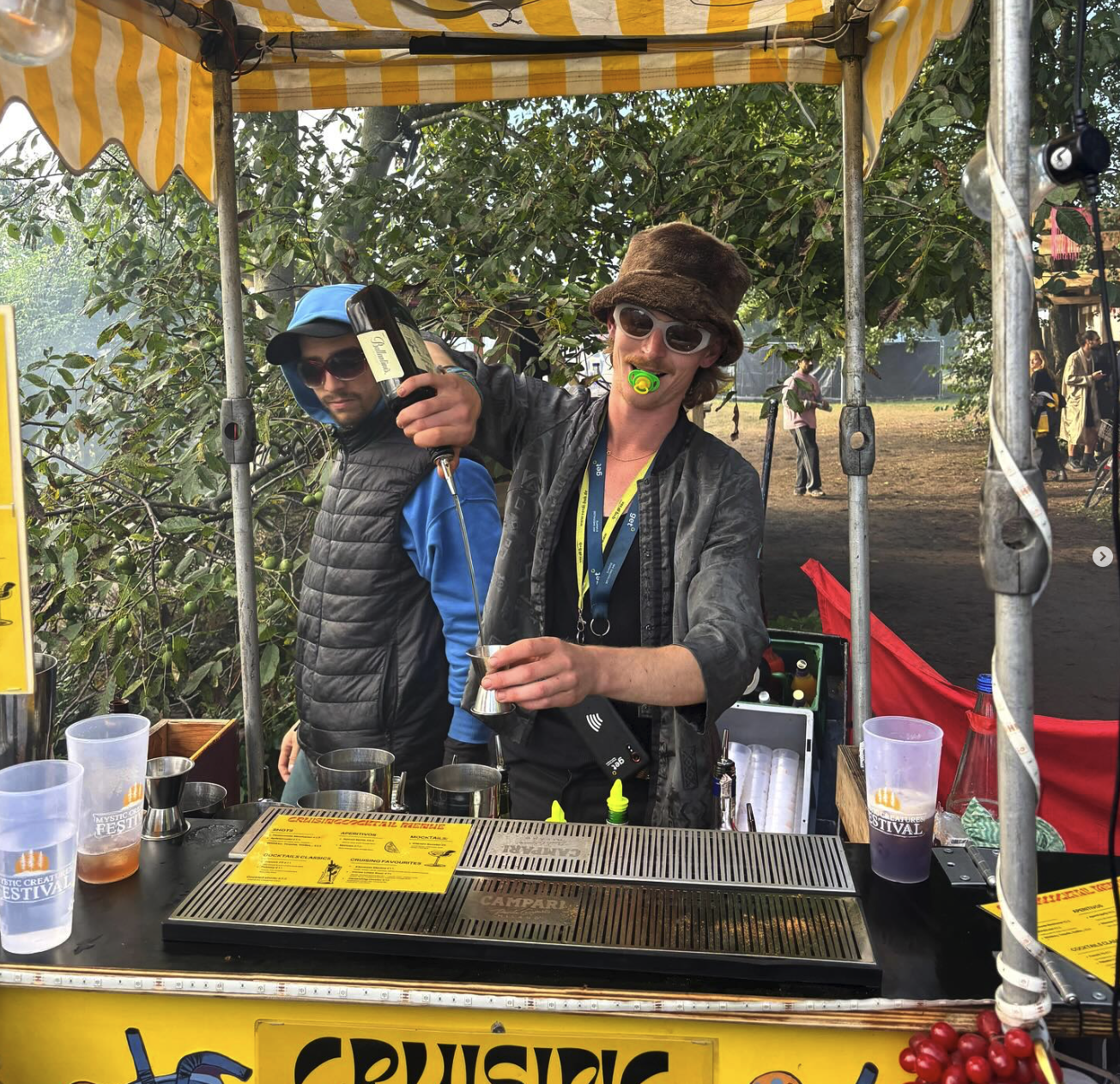 Zwei Bartender schenken unter der gelb-weiß gestreiften Markise des CruisingCocktails-Carts auf einem Festival Drinks aus