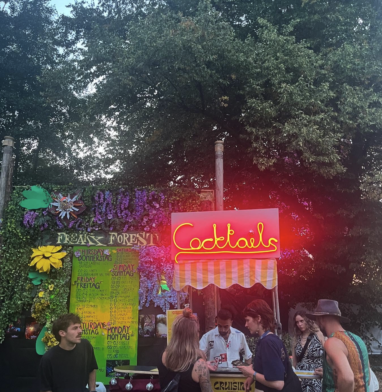 Leuchtend pinkes „Cocktails“-Neonschild über dem Cart bei einem abendlichen Walddancefloor-Event