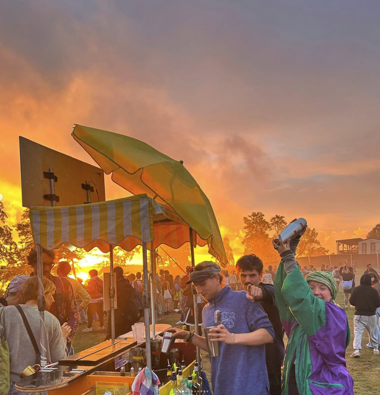 CruisingCocktails-Cart mit grünem Sonnenschirm vor einem dramatischen orangefarbenen Sonnenuntergang, umringt von Festivalgästen